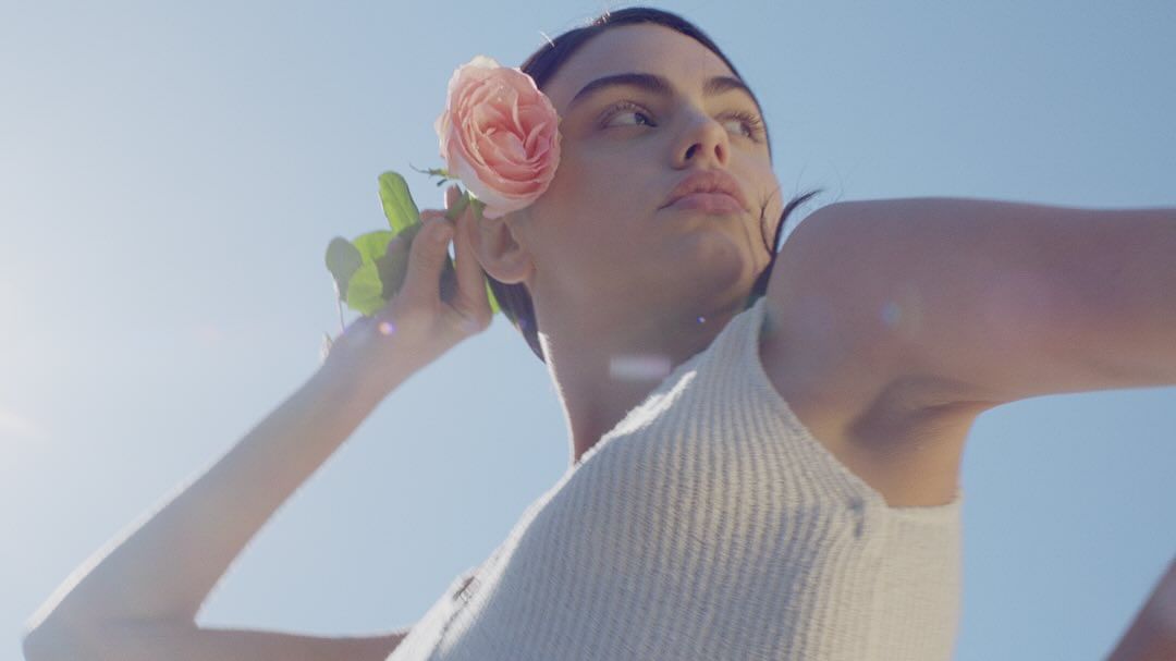 A young adult female with a neutral facial expression stands outside with a rose flower near her ear against a clear blue sky.