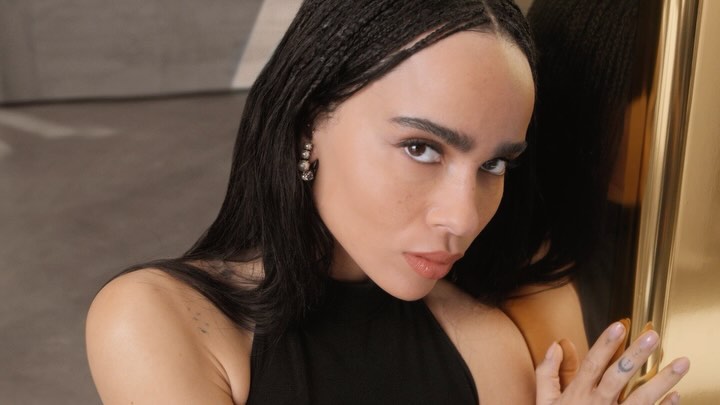 Close-up portrait of Zoë Kravitz with dark braided hair wearing a black halter top, touching a gold framed mirror.
