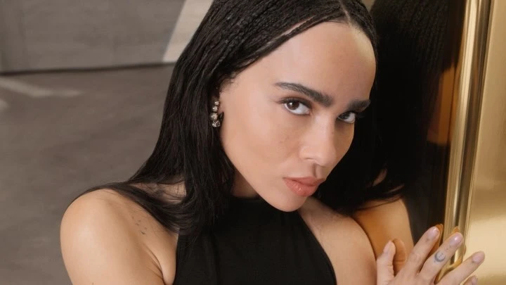 Close-up portrait of Zoë Kravitz with dark braided hair wearing a black halter top, touching a gold framed mirror.