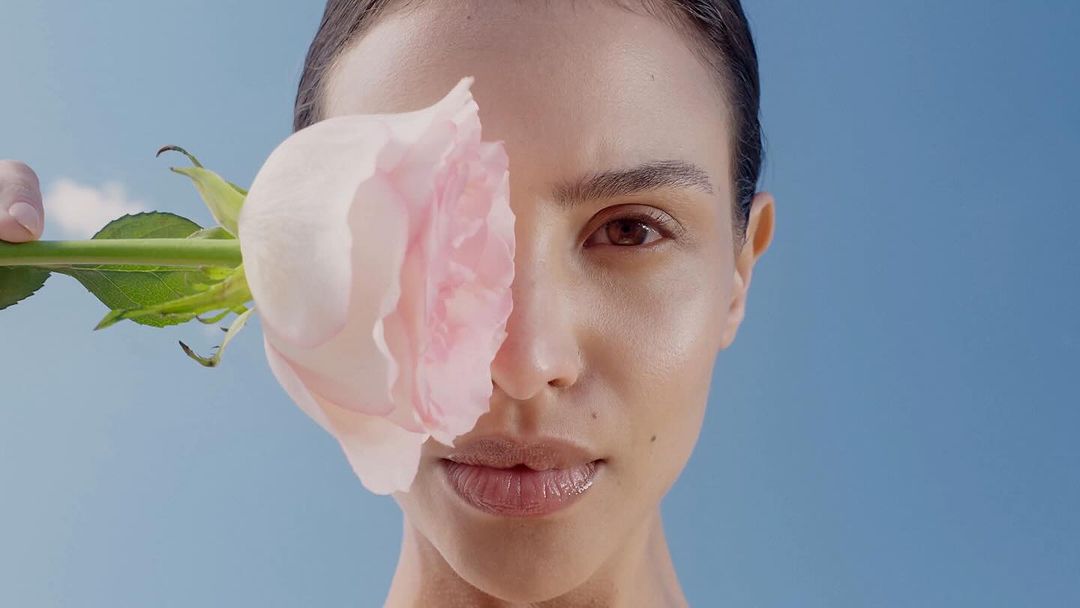 Close-up of a light-skinned woman with a neutral expression holding a pink rose covering one eye against a soft blue background.