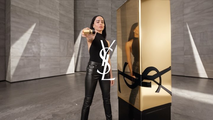 Fashion shot of an adult woman holding a gold cosmetic product next to a gold reflective column with a grey stone background.