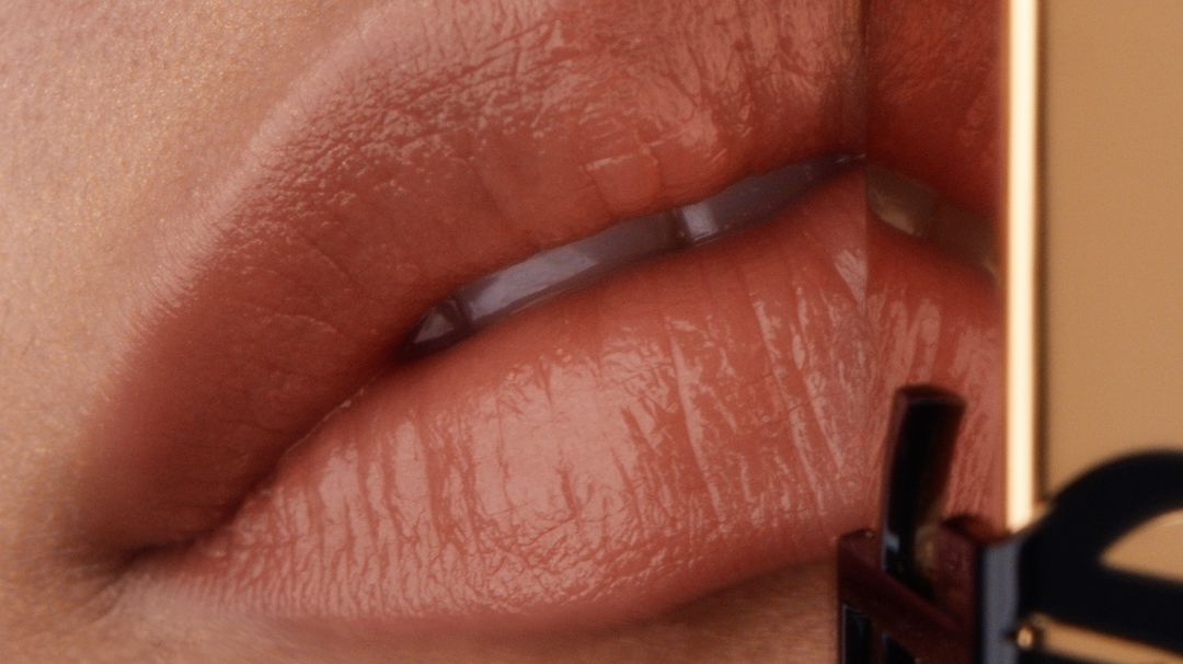 Close-up macro shot of glossy nude lips wearing lipstick beside a gold cosmetic product case by Arthur Valverde.