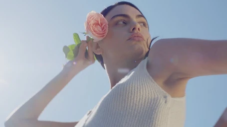 Close-up portrait of a woman holding a pink rose near her ear against a bright blue sky background.