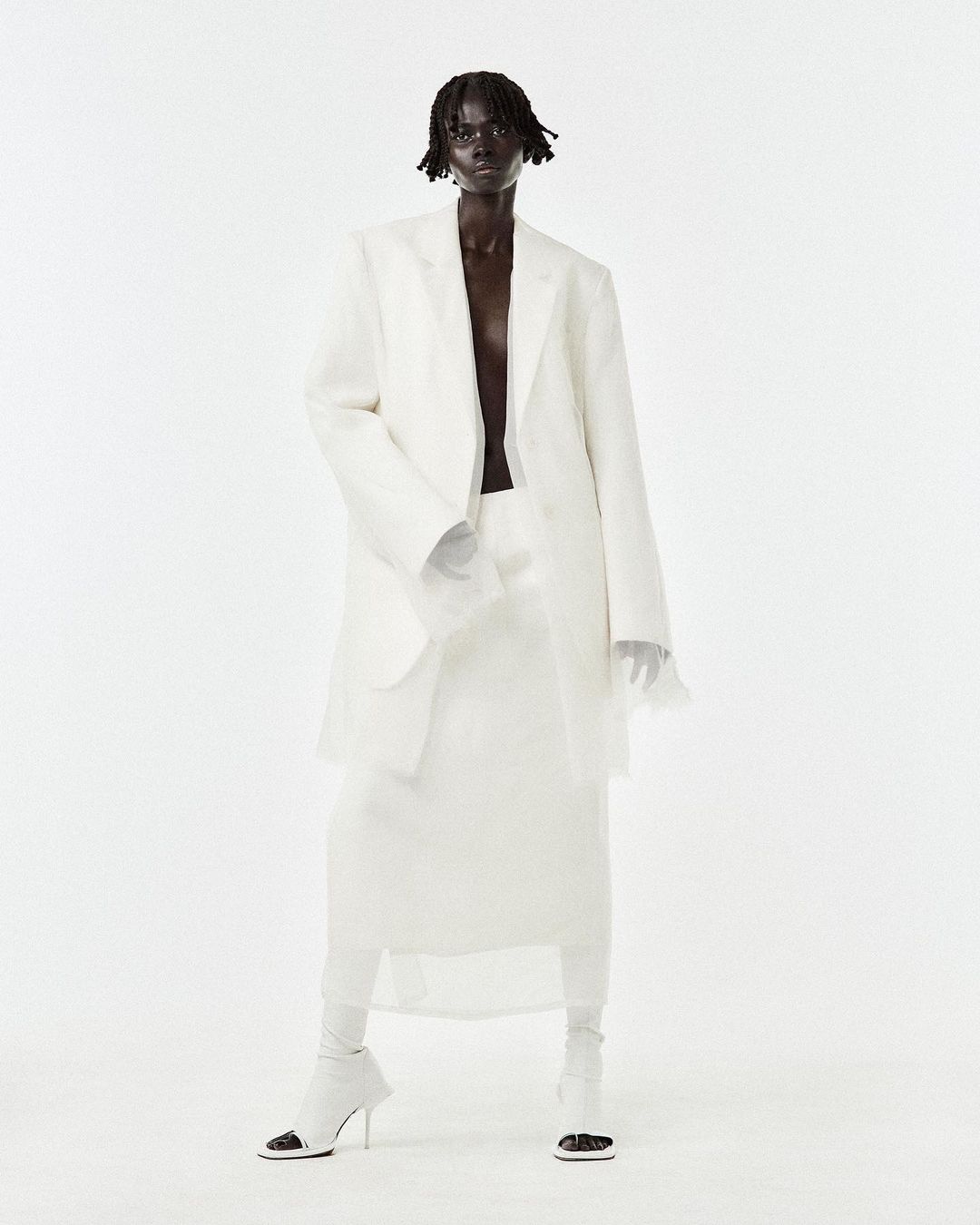 Full-length shot of a single, dark-skinned adult woman in a loose, all-white suit and high-heeled shoes, against a bright white backdrop.