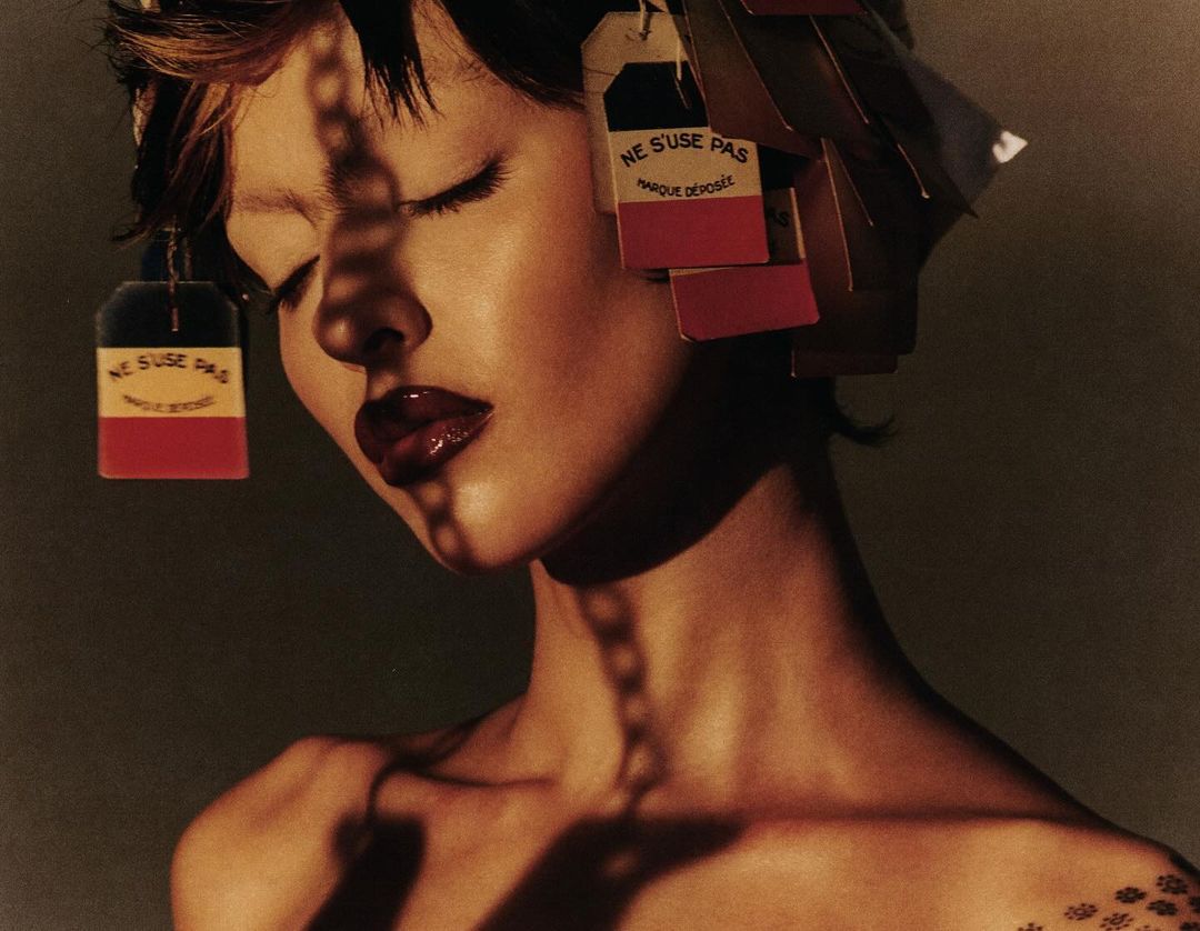 Close-up shot of an adult female with closed eyes and dark glossy lips wearing multiple tags in her hair, with shadow on her face.