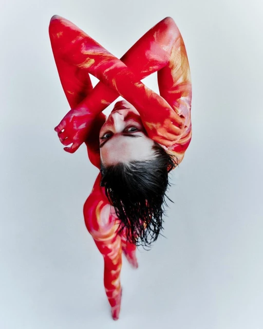 Woman covered in red and orange body paint is standing against a white backdrop in a top-down angle and arms crossed.