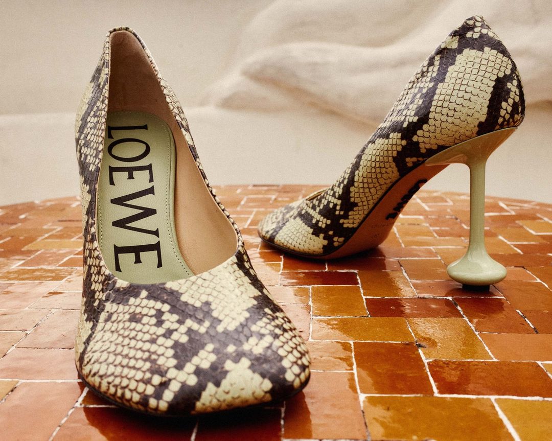 Two Loewe heeled pumps with a snake-skin pattern rest on a geometric terra-cotta surface, against an out-of-focus white wall.