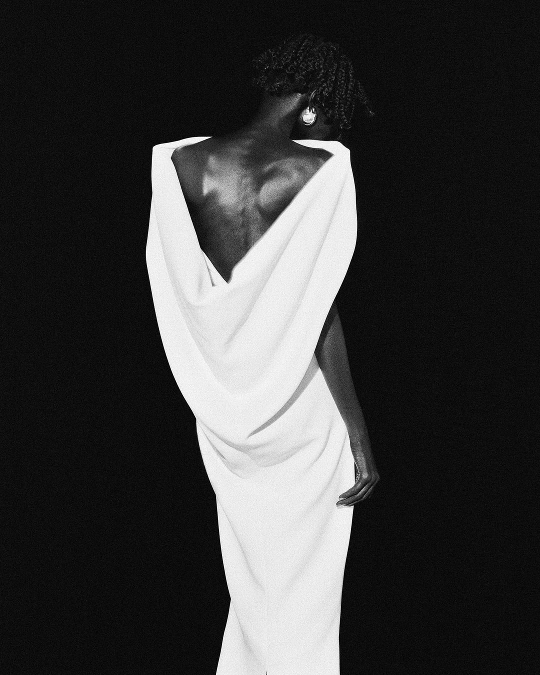 Elegant studio portrait of an adult woman with dark hair wearing a long white dress, with a black backdrop creating a striking contrast.