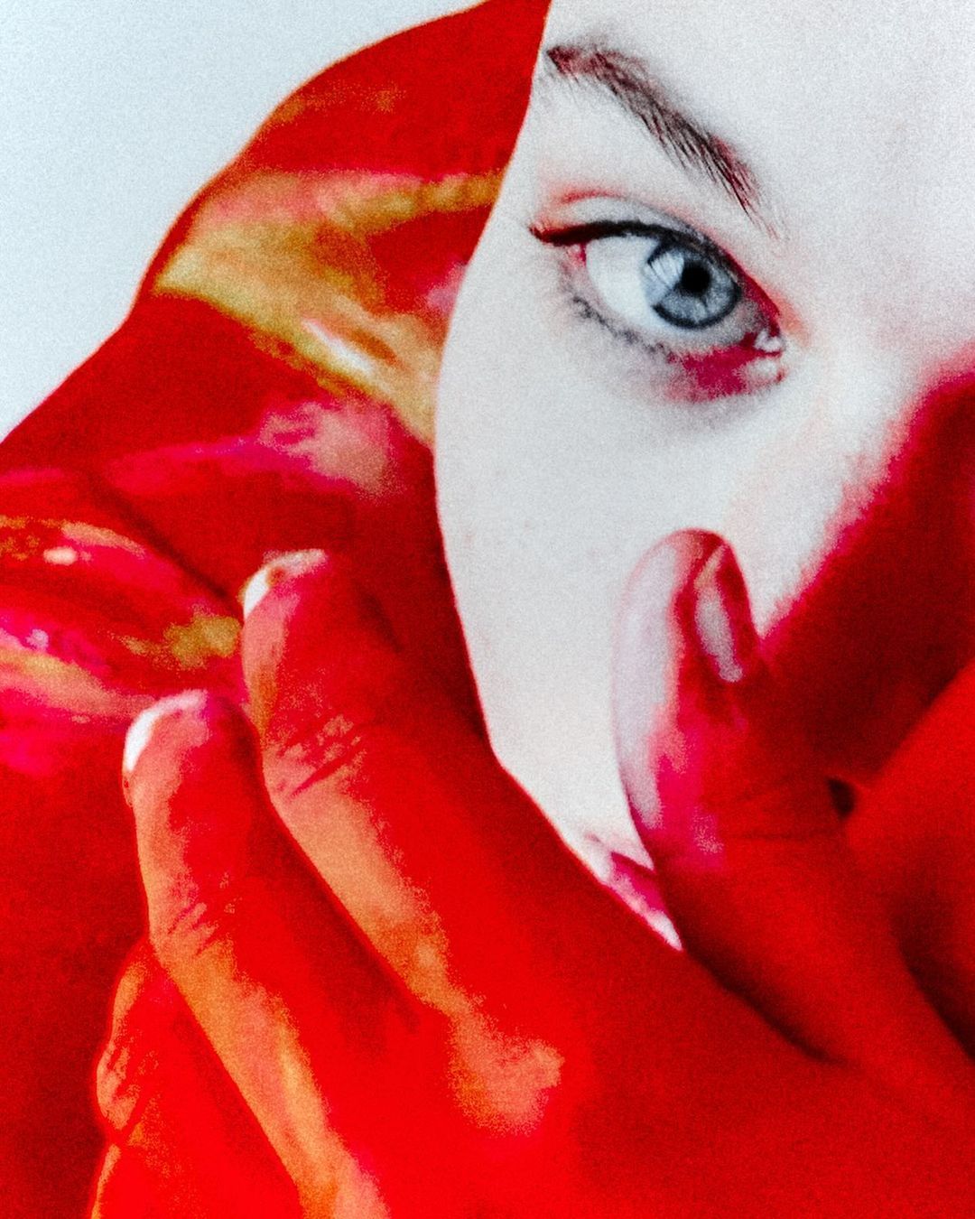 Close-up of a woman's face with fair skin and blue eyes, partially obscured by a red-toned hand. The image has a soft, filtered appearance.