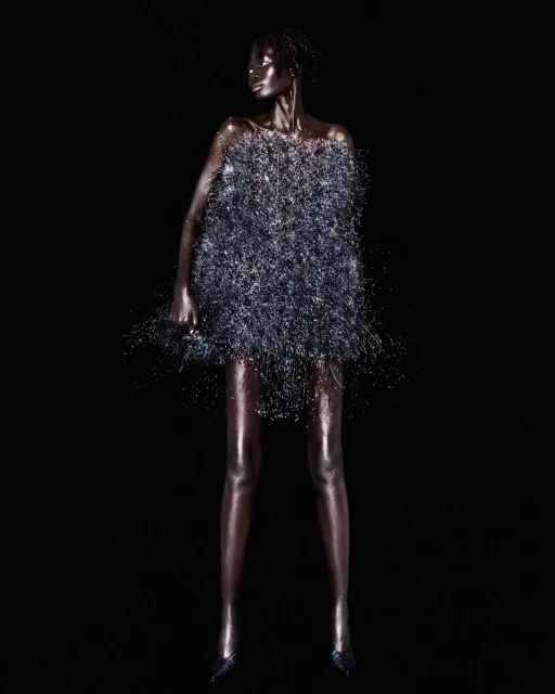 Full-length studio shot of a bronze-toned model wearing a silver metallic dress, posing against a dark black background with high fashion.