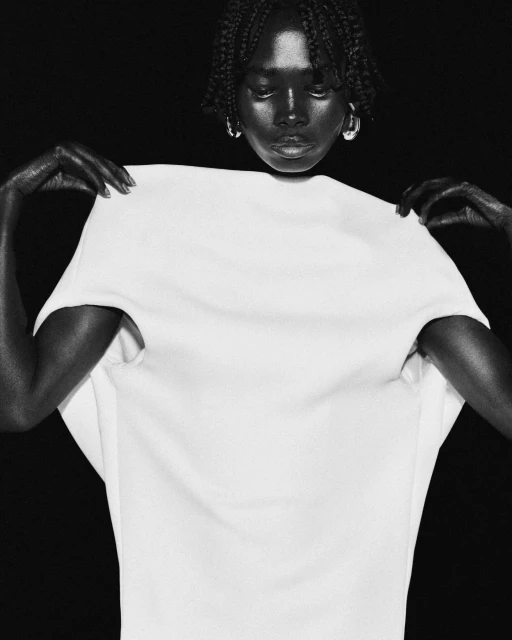 Black and white portrait of an African-descent woman holding up a white top against a black background, showcasing the garment.