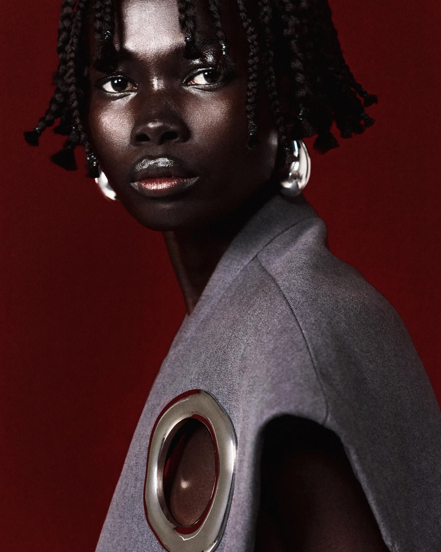 Close-up portrait of a dark-skinned Black woman with braided hair wearing silver hoop earrings and a gray jacket against a red backdrop.