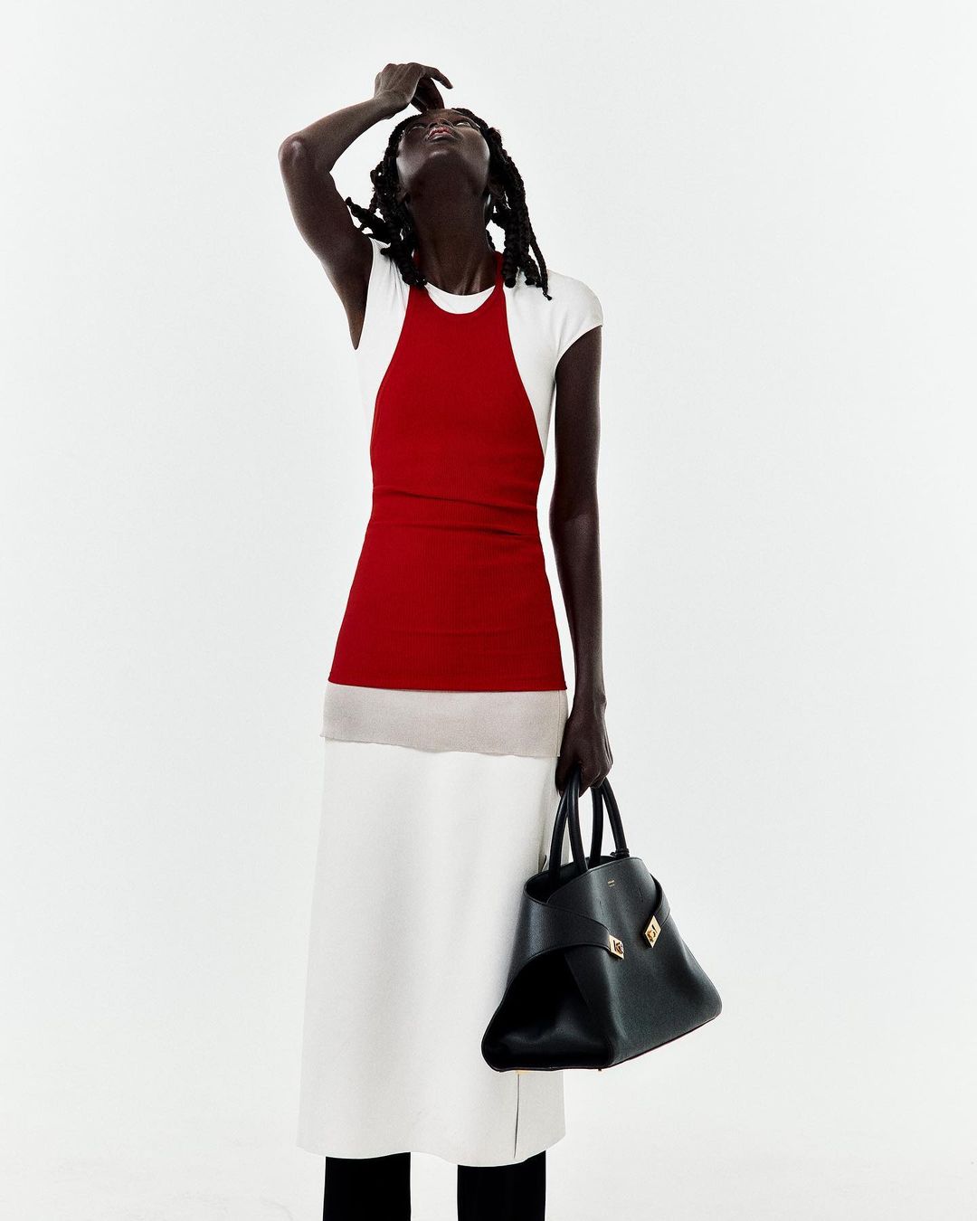 An elegant Black woman in a studio poses in a white and red dress, holding a black leather bag. The bright background gives it a fashion editorial look.
