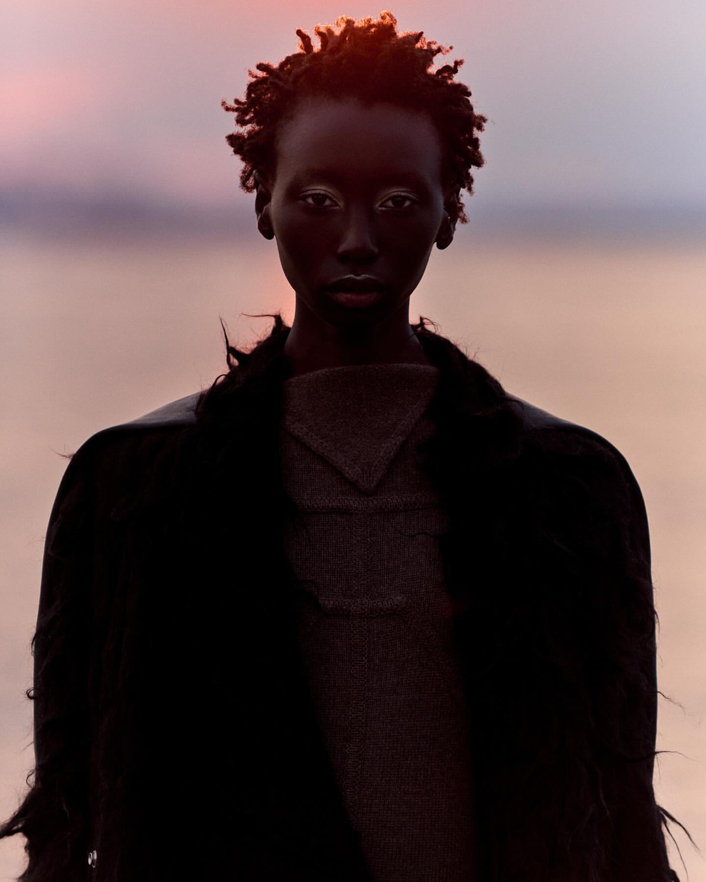 Portrait of a Black female model with white eye makeup, wearing a dark coat and top, standing outdoors against a soft sunset background.