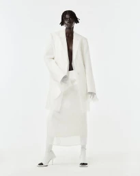 Model wearing an oversized white blazer and matching skirt with heeled boots against a seamless white studio background.