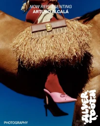 Person riding a horse while holding a brown straw fringed handbag with a gold B logo against a solid blue sky background