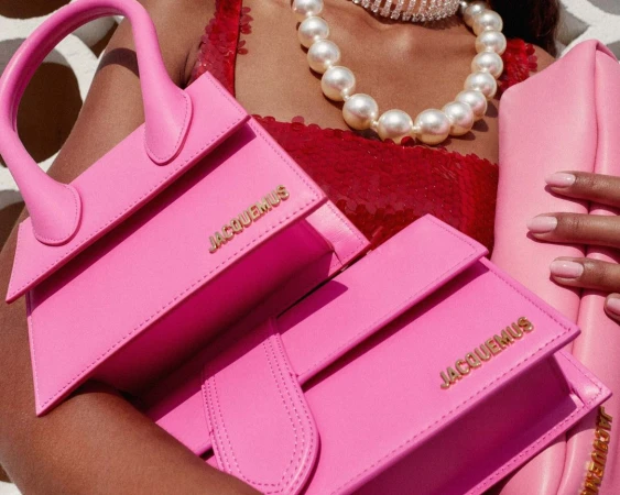 Close-up portrait of a person holding three pink Jacquemus handbags, wearing a pearl necklace and red sequin top.