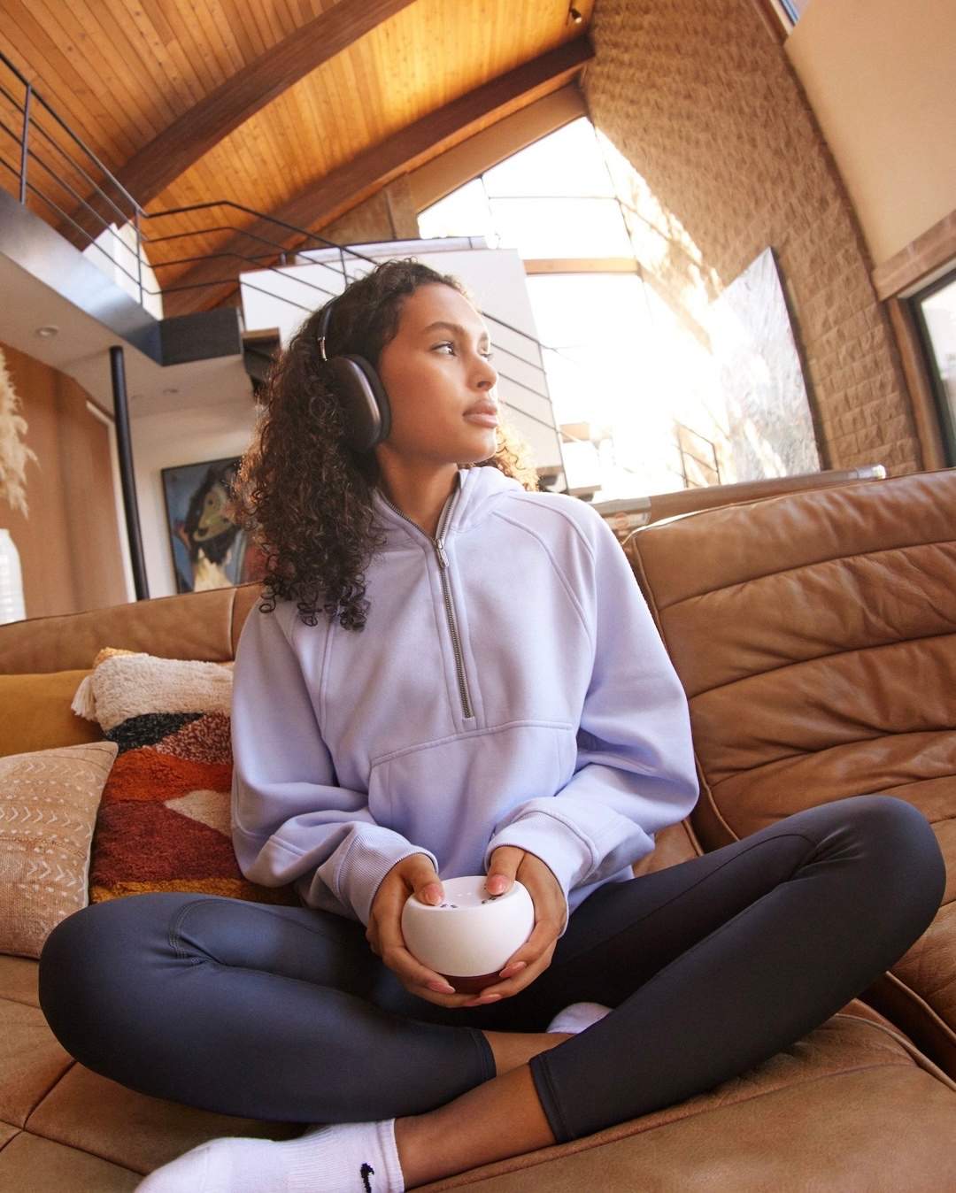 Young woman with headphones, wearing a blue hoodie and leggings, relaxing on a brown sofa in a brightly lit, modern home interior.