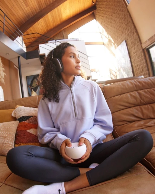Young woman with headphones, wearing a blue hoodie and leggings, relaxing on a brown sofa in a brightly lit, modern home interior.