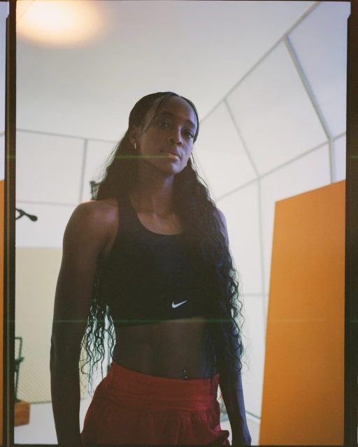 Athletic woman in sports bra and shorts stands indoors with a determined expression, showcasing fitness and healthy lifestyle, with orange accents.