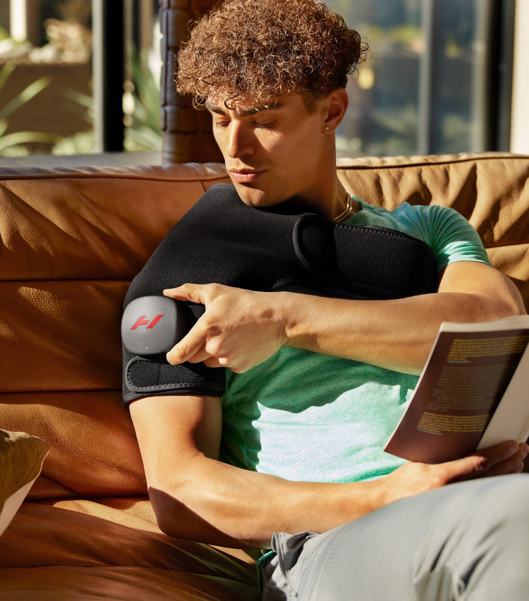 Adult male sits on a couch wearing a shoulder wrap and reading a book; natural light from a window.
