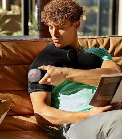 Adult male sits on a couch wearing a shoulder wrap and reading a book; natural light from a window.