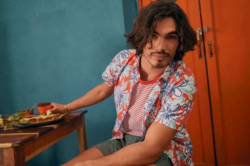 Man in floral shirt sits at a table with food and drinks in front of colorful walls, capturing a casual dining scene with vibrant hues.