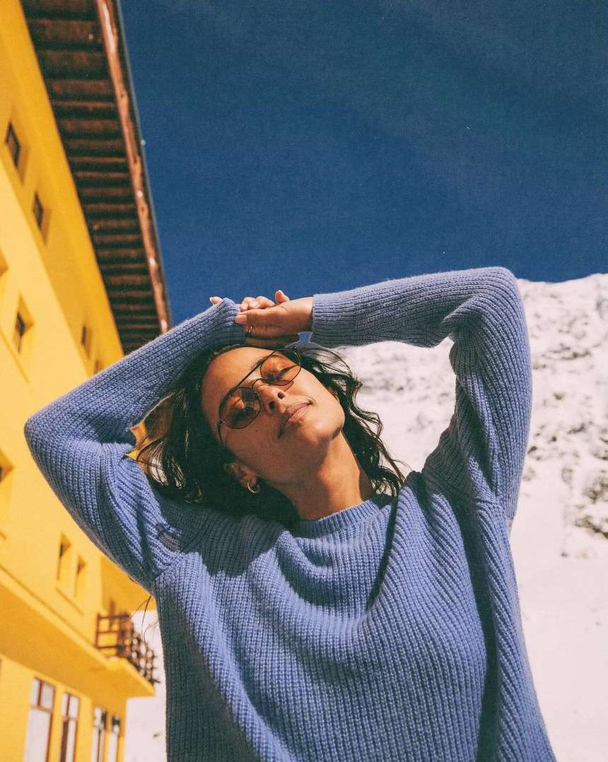 Woman in blue sweater poses in front of yellow building, blue sky, and snow-covered mountain on a bright day with sunglasses.
