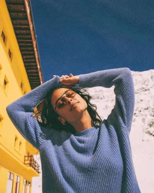 Woman in blue sweater poses in front of yellow building, blue sky, and snow-covered mountain on a bright day with sunglasses.