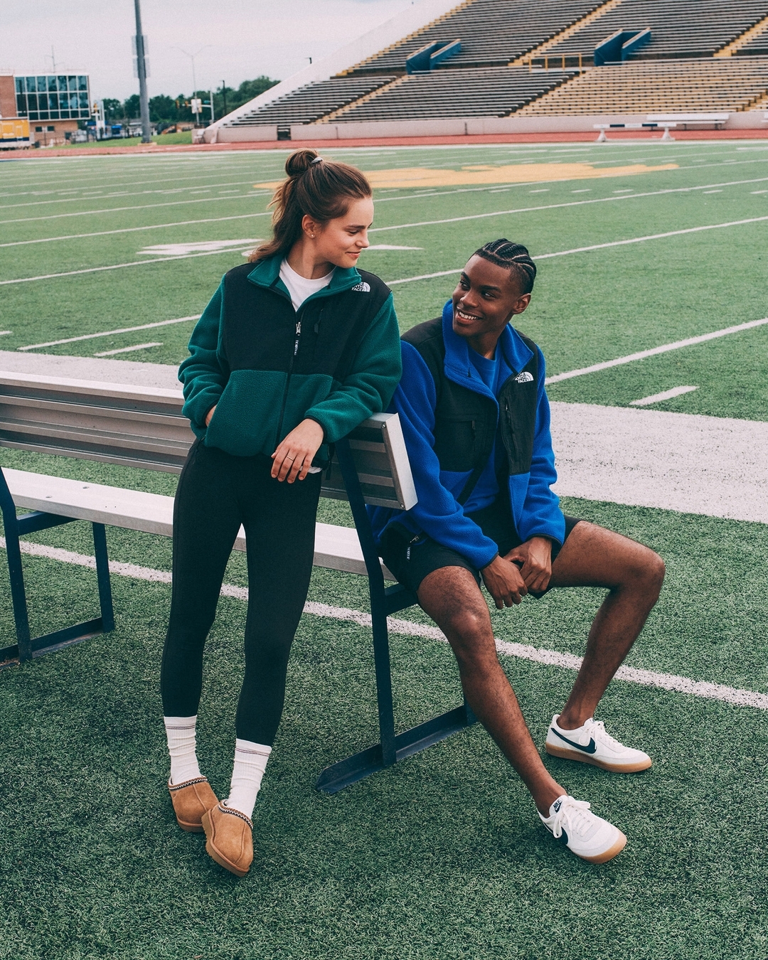 Two people, an adult woman standing and an adult man sitting, wear North Face fleece jackets in an outdoor setting on a football field.