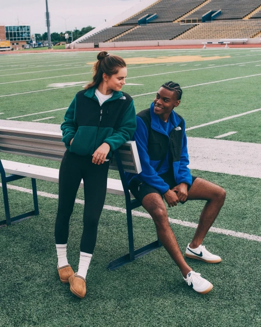 Two people, an adult woman standing and an adult man sitting, wear North Face fleece jackets in an outdoor setting on a football field.