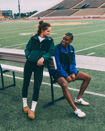 Two people, an adult woman standing and an adult man sitting, wear North Face fleece jackets in an outdoor setting on a football field.