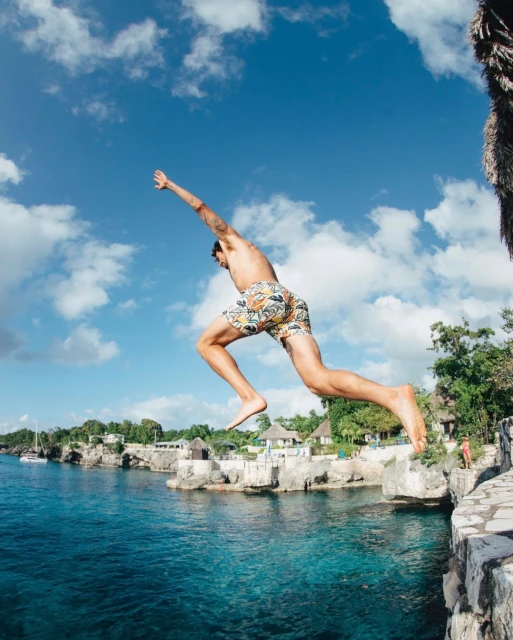 Adult male jumps off a stone cliff into turquoise ocean water, a tropical coastline is visible, with a bright blue sky.