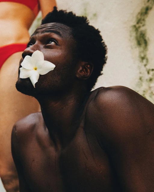 Portrait of an African American man with a white flower near his mouth, looking up, with another person in a red top in the background.