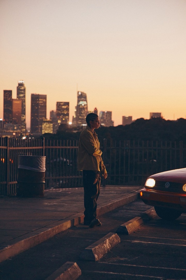 Man stands in urban setting near car with Los Angeles skyline in the background at dusk. Tranquil city scene with warm colors.