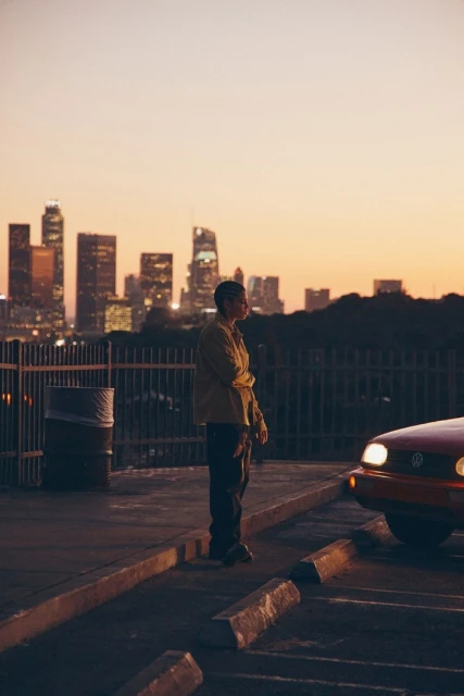 Man stands in urban setting near car with Los Angeles skyline in the background at dusk. Tranquil city scene with warm colors.