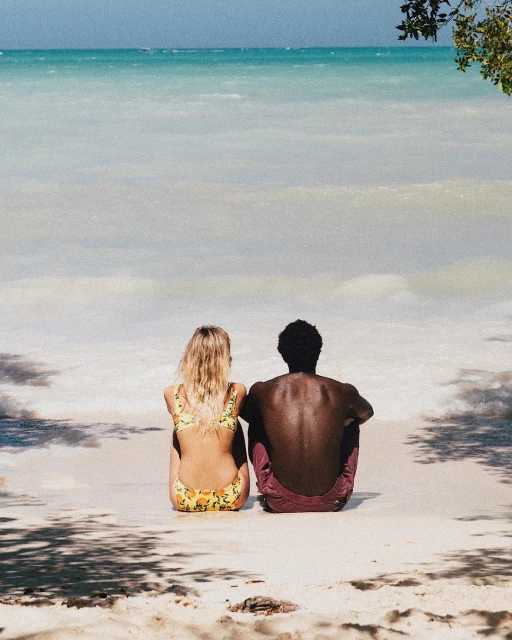 Couple relaxing on beach: Blond woman in bikini and Black man in shorts sit on sand near ocean, framed by shadows and turquoise water.