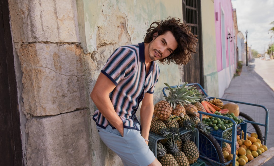 Adult man leans against a stone wall, smiling, wearing a striped shirt and shorts, with a fruit cart in the background on a sunny street.