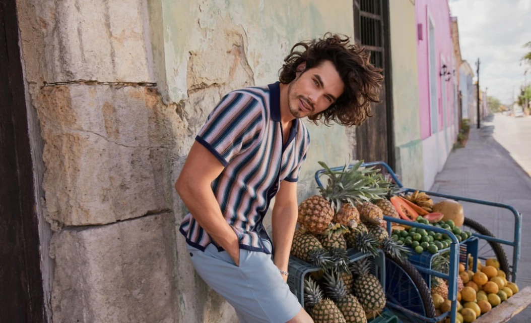 Adult man leans against a stone wall, smiling, wearing a striped shirt and shorts, with a fruit cart in the background on a sunny street.