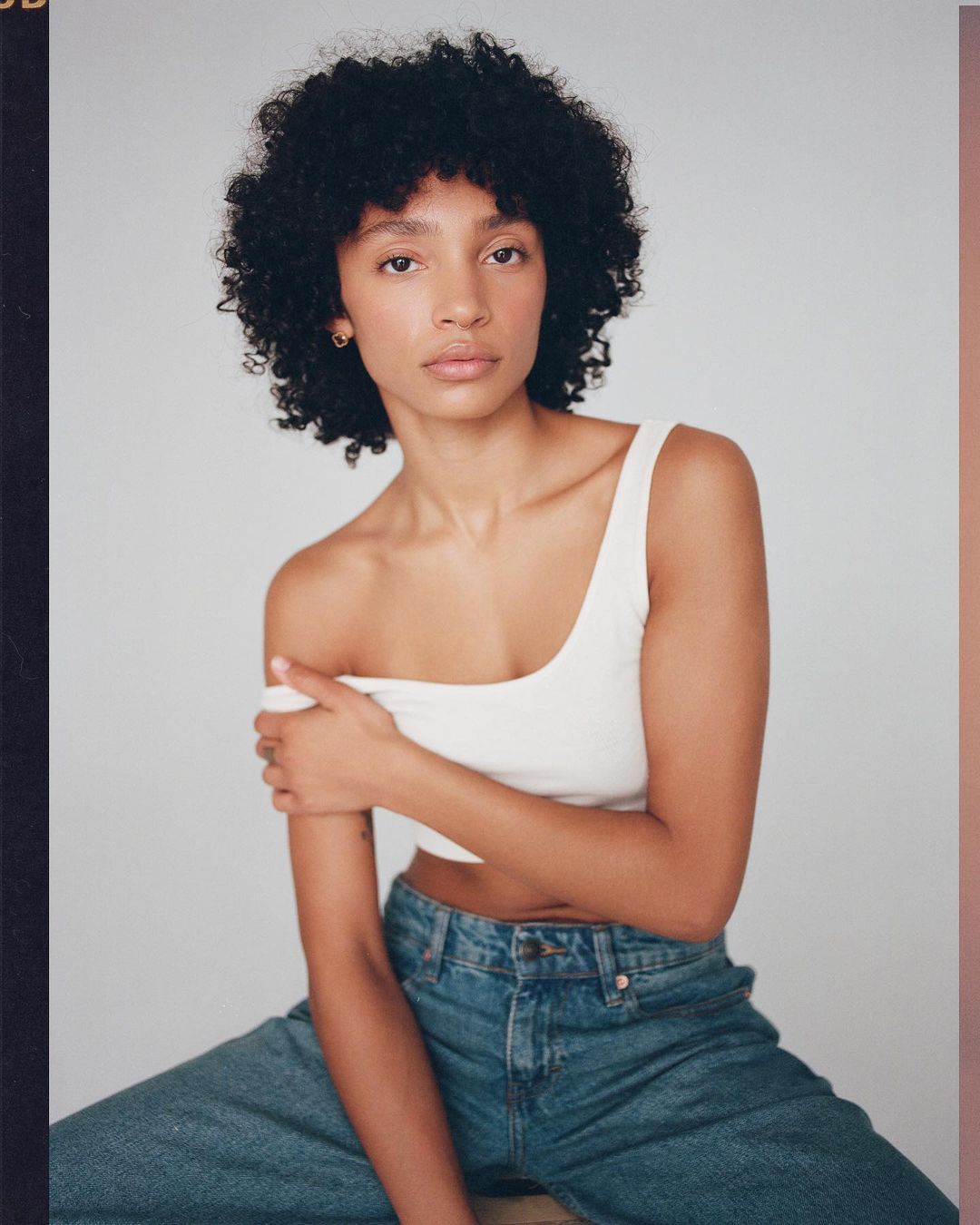 A young woman with curly hair is seated, wearing a white top and jeans, in a studio setting with soft lighting, posing for a portrait.