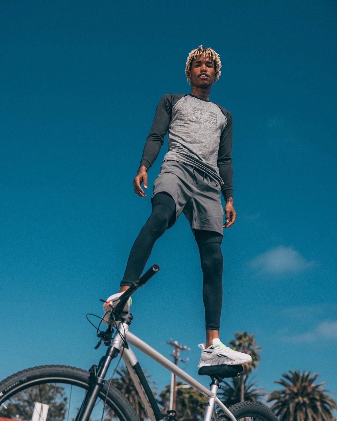 Athletic adult male stands confidently with one leg on a silver and black mountain bike outdoors against a blue sky backdrop.