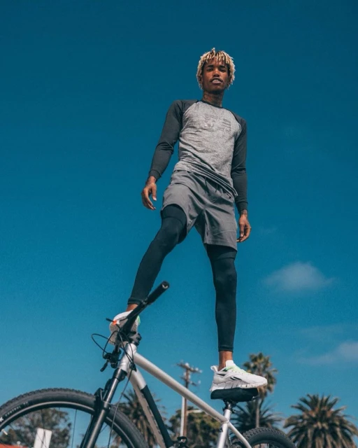 Athletic adult male stands confidently with one leg on a silver and black mountain bike outdoors against a blue sky backdrop.