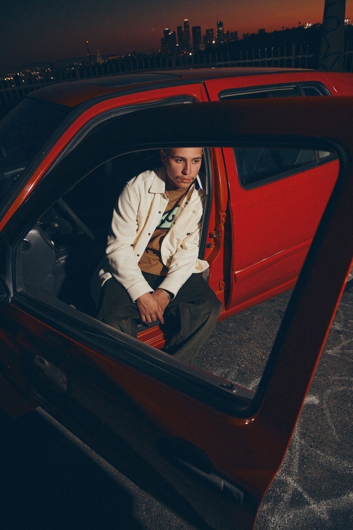 An adult sits in a red car with the door open at dusk, a city skyline behind. The individual wears a cream jacket and dark pants.