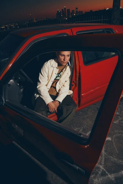 An adult sits in a red car with the door open at dusk, a city skyline behind. The individual wears a cream jacket and dark pants.
