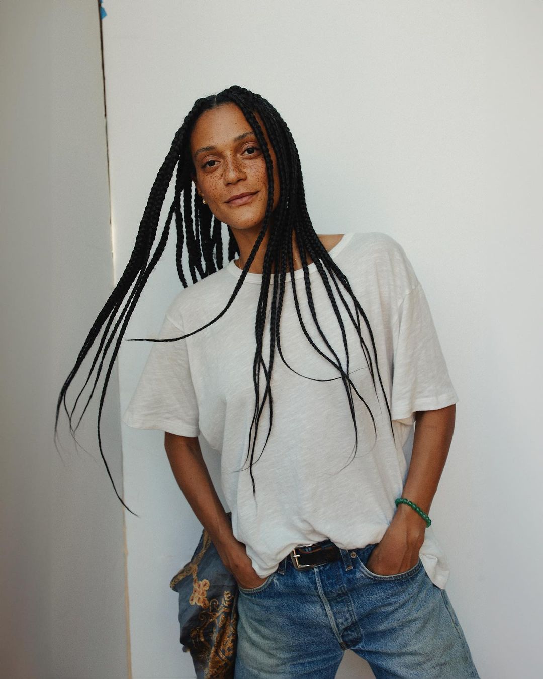 Woman with long braids poses indoors wearing a white t-shirt and jeans, casting a relaxed and casual vibe against a neutral backdrop.