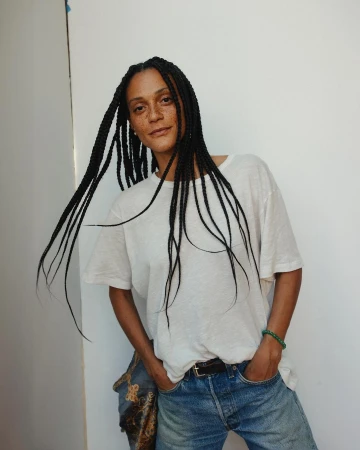 Woman with long braids poses indoors wearing a white t-shirt and jeans, casting a relaxed and casual vibe against a neutral backdrop.