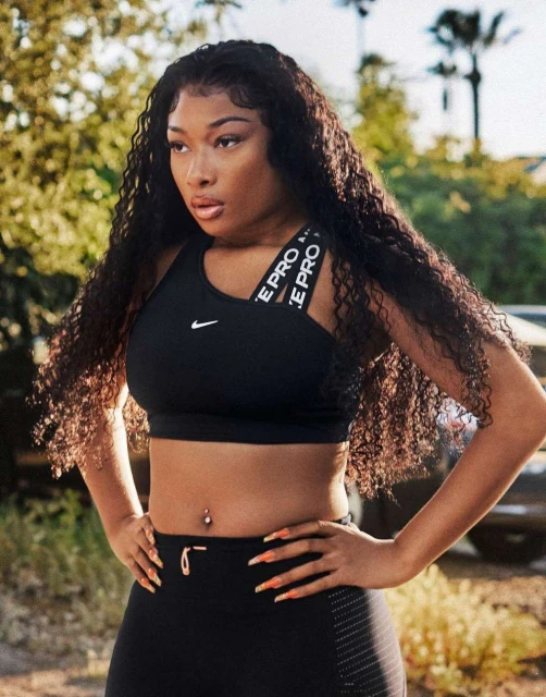 Adult woman in black sports bra and leggings stands outdoors with hands on hips. She has long, curly hair and a neutral facial expression.