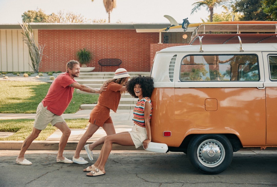Three people push an orange and white vintage camper van on a suburban street with a red brick house and palm trees in the background.