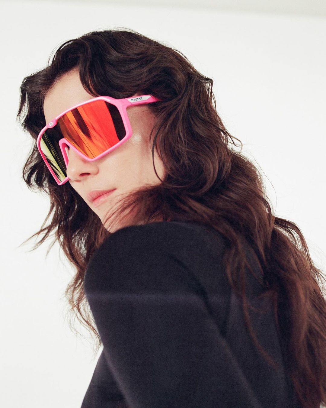 A stylish young woman with long wavy dark hair poses wearing bright pink sunglasses with red lenses against a white background in a portrait.
