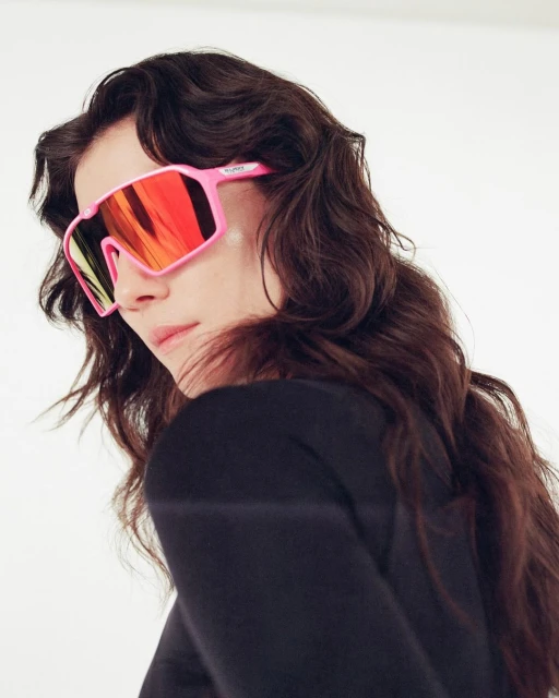 A stylish young woman with long wavy dark hair poses wearing bright pink sunglasses with red lenses against a white background in a portrait.